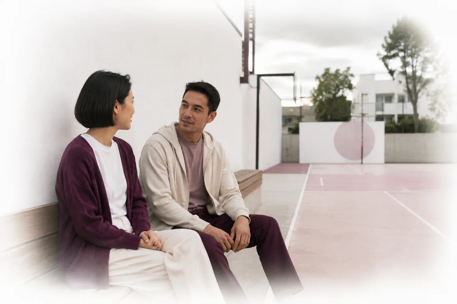 A woman and a man talking face to face on a bench beside a neighbourhood sports court.