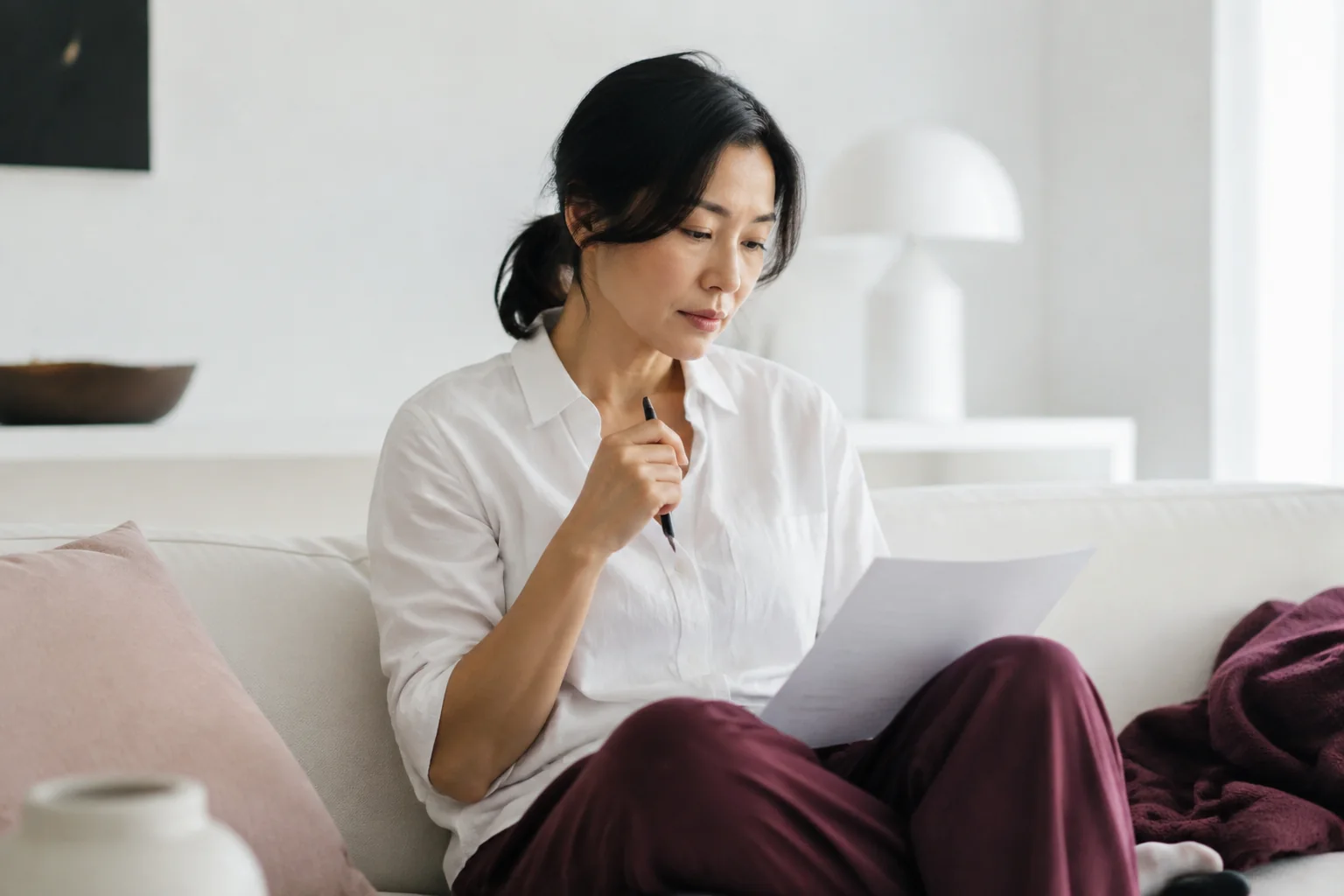 A woman pauses over handwritten pages while sitting cross-legged on a pale sofa.