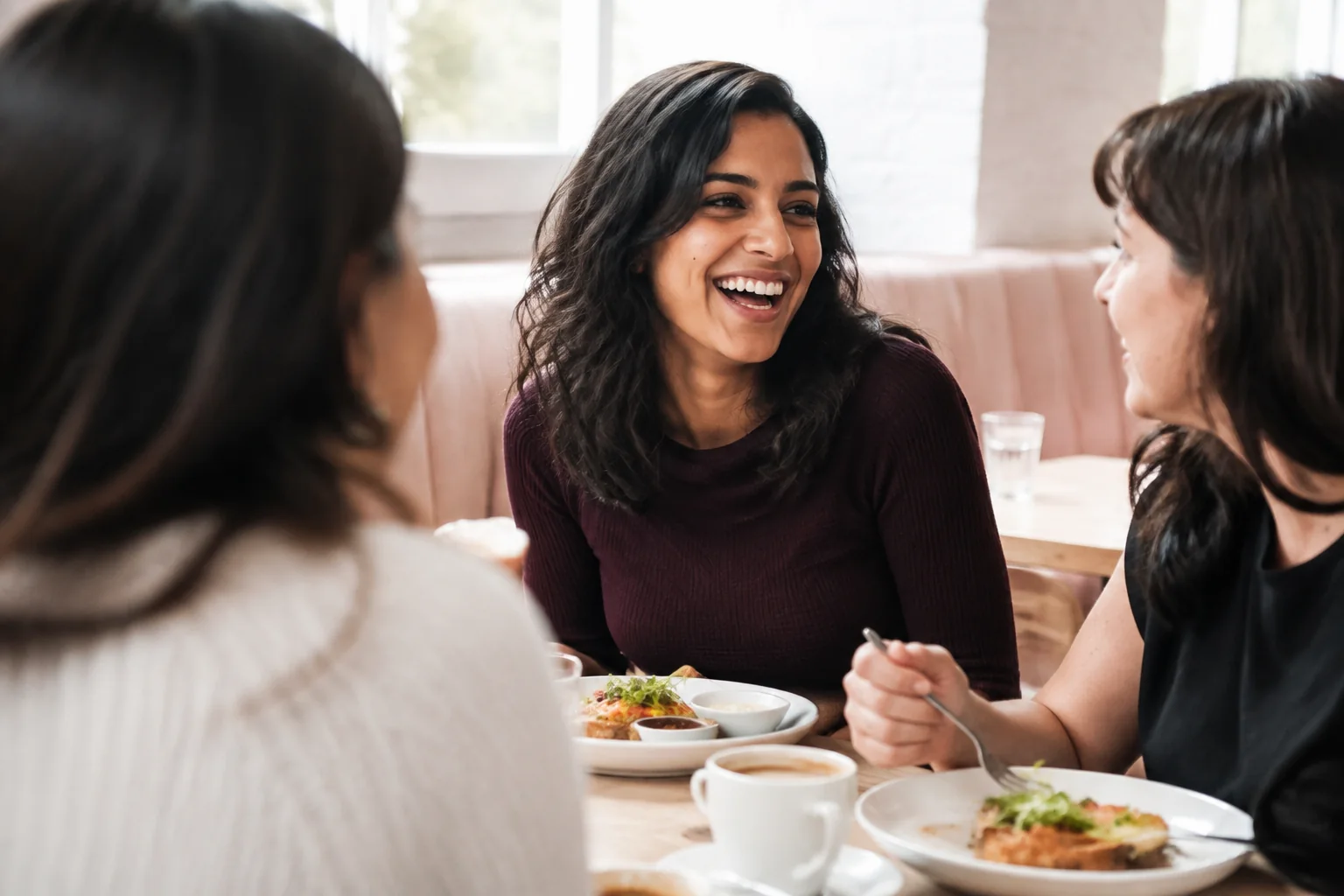 A woman laughing with two friends over breakfast in a light-filled cafe.