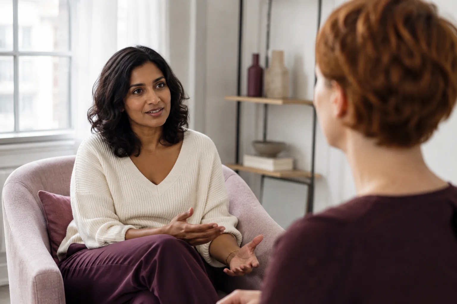 Two friends talking from separate chairs in a light-filled sitting room.