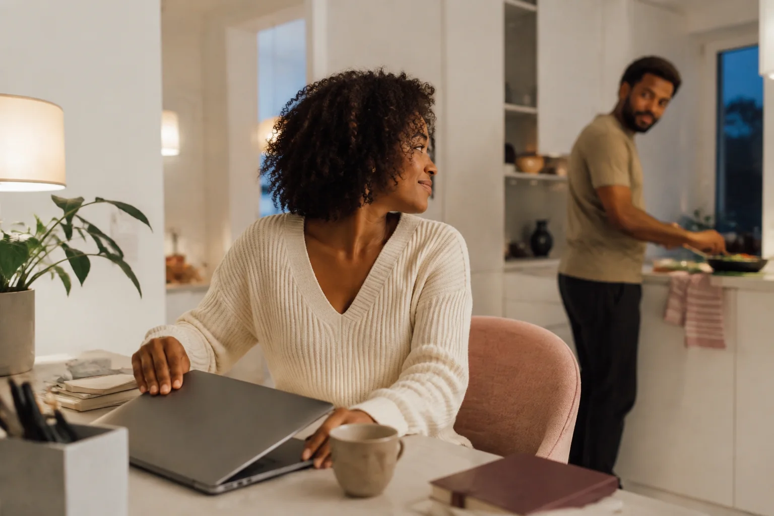 A woman closing her laptop while her partner prepares dinner nearby.