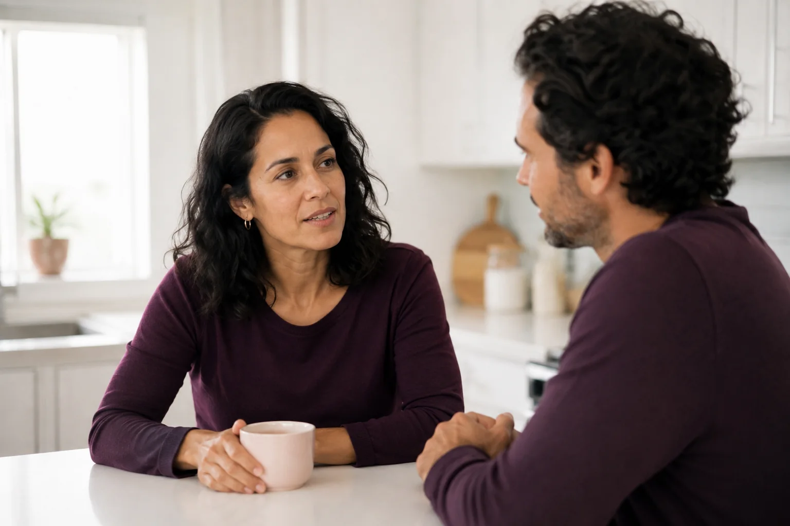 A woman in a plum top talking with her partner at a bright kitchen counter.